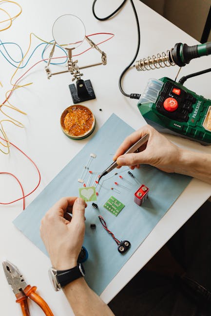 Hands assembling electronic components on a work table with tools and wires.